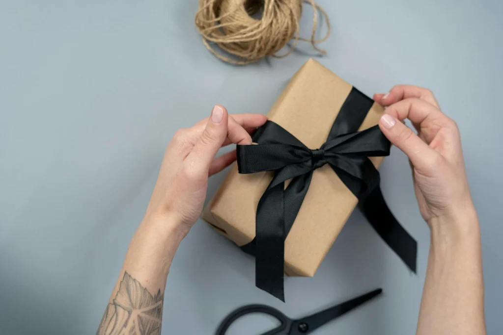 In the picture two hands are holding a bow of a gift package. The ribbon is black and the paper of the package is brown.