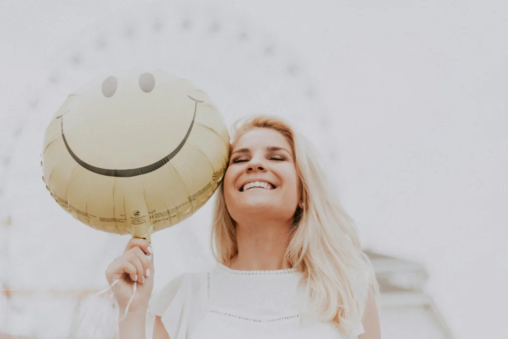 In the picture a smiling girl is holding a yellow balloon with a smiley face drawn on it in her hand.