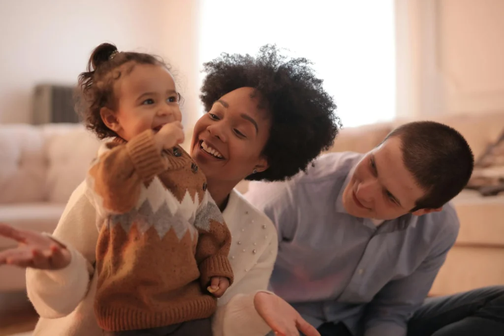 The picture shows a family with mother, father and daughter. The smiling parents are watching the little girl nibbling on a piece of cookie.