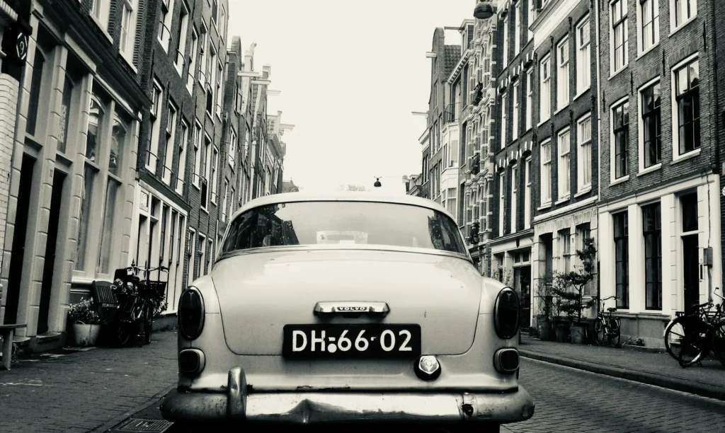 The image shows the back of an old vintage Volvo parked in a narrow street with brick Anglo-Saxon house fronts on both sides. The photo is in black and white.