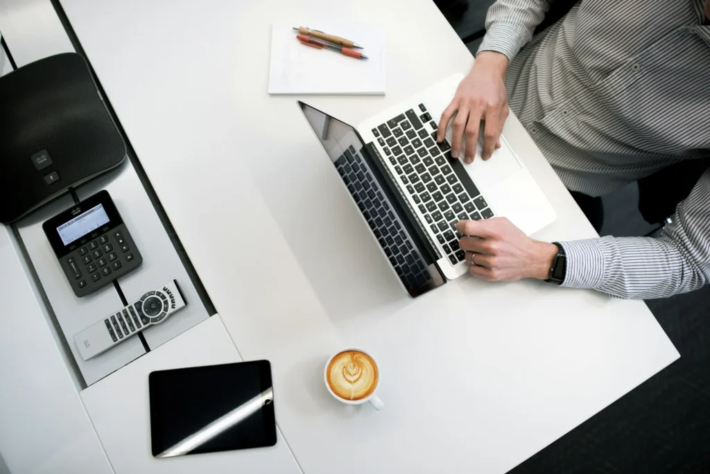 In the image a man in a striped shirt works on a laptop placed on a white desk. On the desk there are also: a notebook and 2 pens next to the PC, a cappuccino and a tablet; on the left a remote control, a speaker and a monitor with a keyboard.