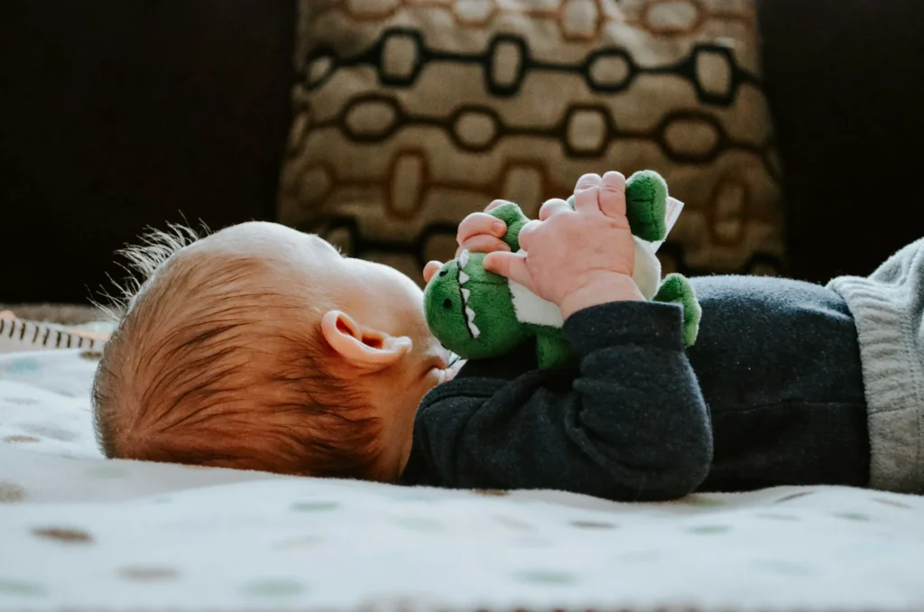 Nell'immagine un bimbo è sdraiato su un letto e tiene nelle mani un peluche a forma di drago verde.