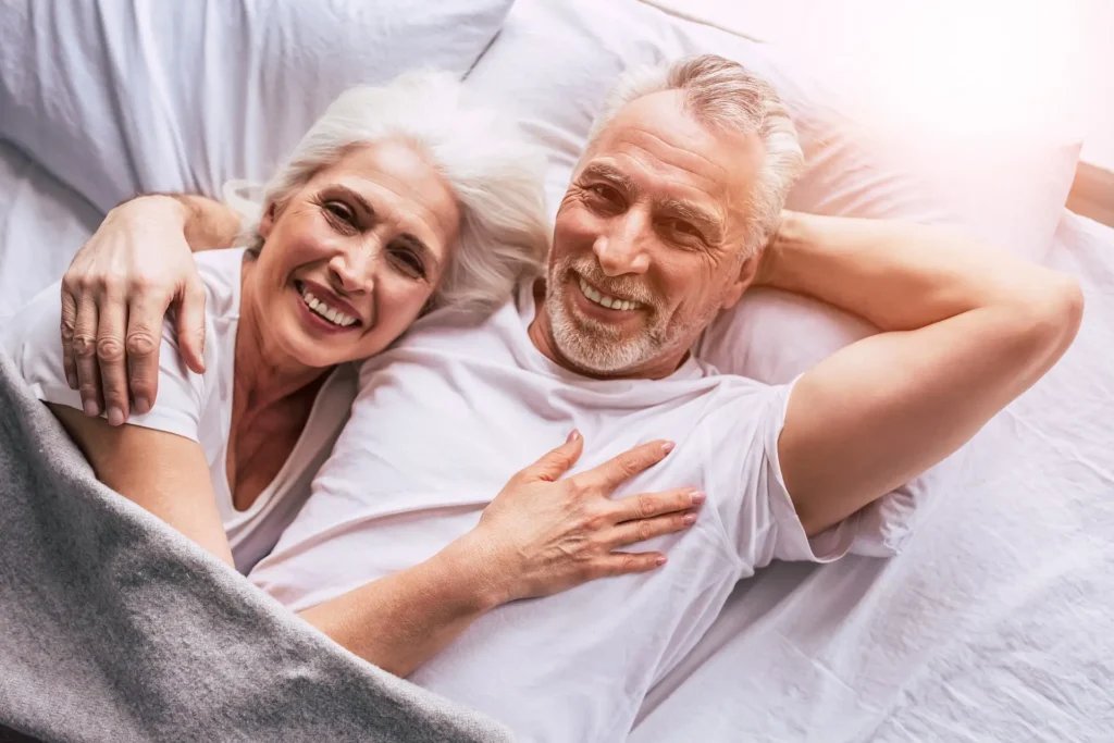 In the picture a senior couple lying on a bed hugs and smiles