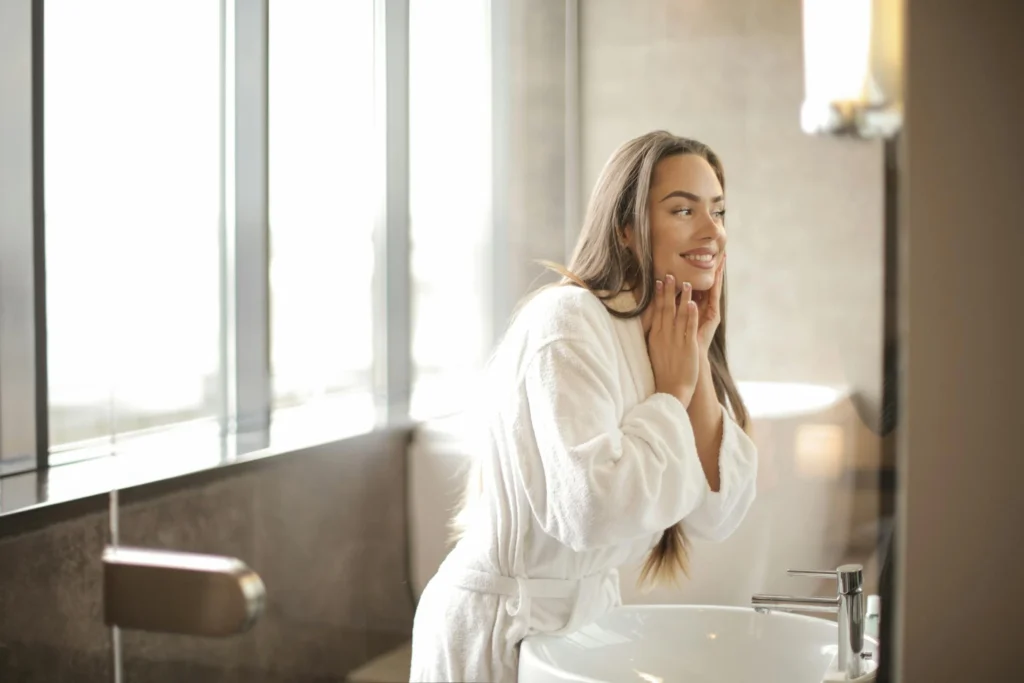 In the picture a girl in a bathrobe smiles while looking at herself in the mirror.